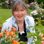 Die Fotografin Nathalie Dautel inmitten blühender Rosen. Direkt vor ihr ein Rosenstrauch in Orangetönen.