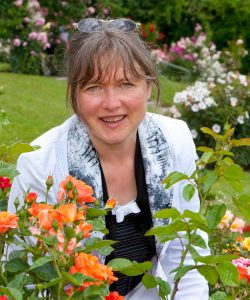 Die Fotografin Nathalie Dautel inmitten blühender Rosen. Direkt vor ihr ein Rosenstrauch in Orangetönen.